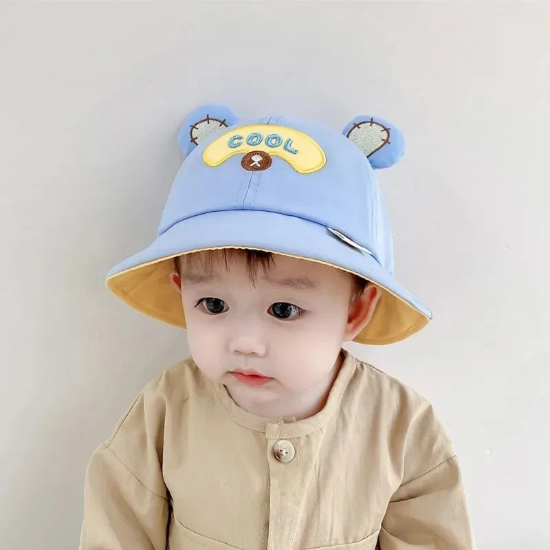 Baby wearing a green frog bucket hat, smiling, showcasing adorable sun protection. Toddler summer fashion.