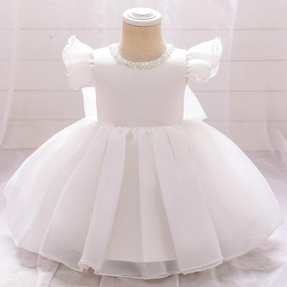 Toddler girl wearing white baptismal princess dress with large bow and delicate beading, full length.