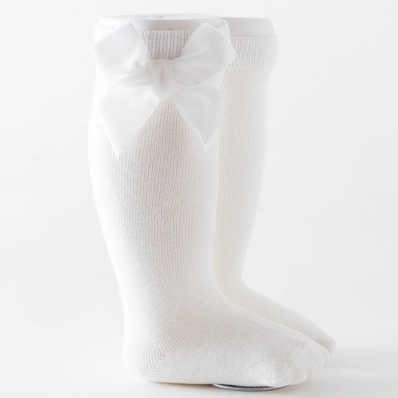 Little girl's feet wearing white knee-high socks with a large bow, on a wooden floor.
