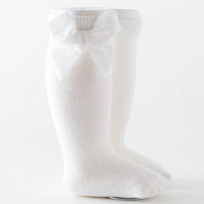 Little girl's feet wearing white knee-high socks with a large bow, on a wooden floor.