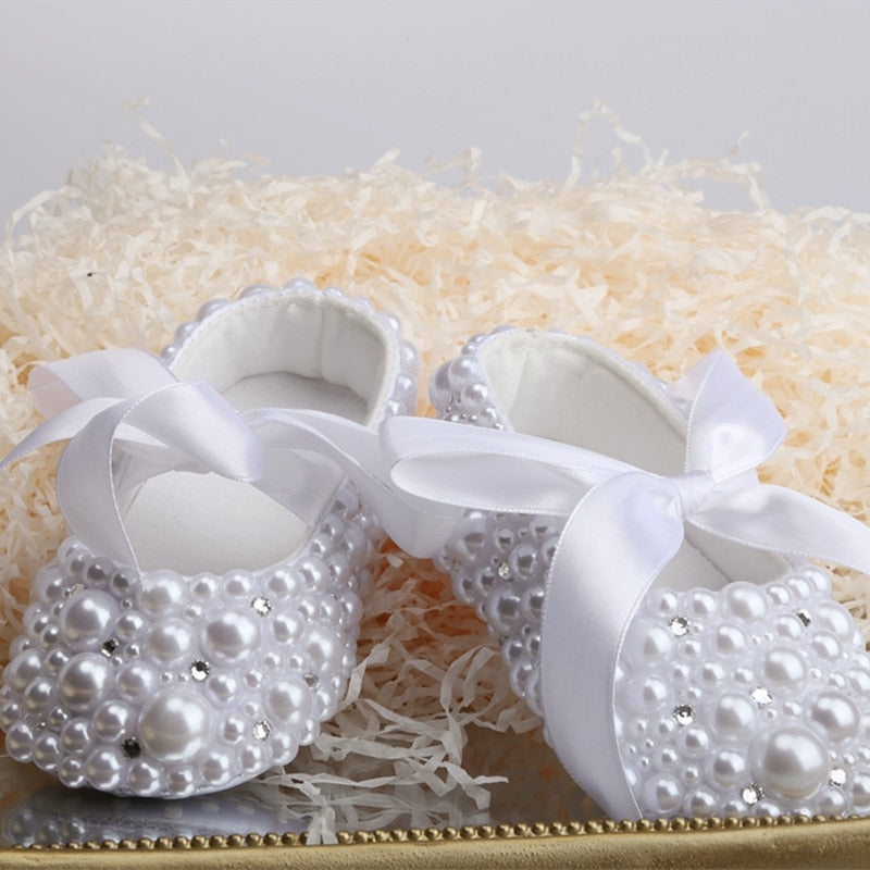 Close-up of baby girl's pearl and ribbon first walker shoes, perfect for special occasions.
