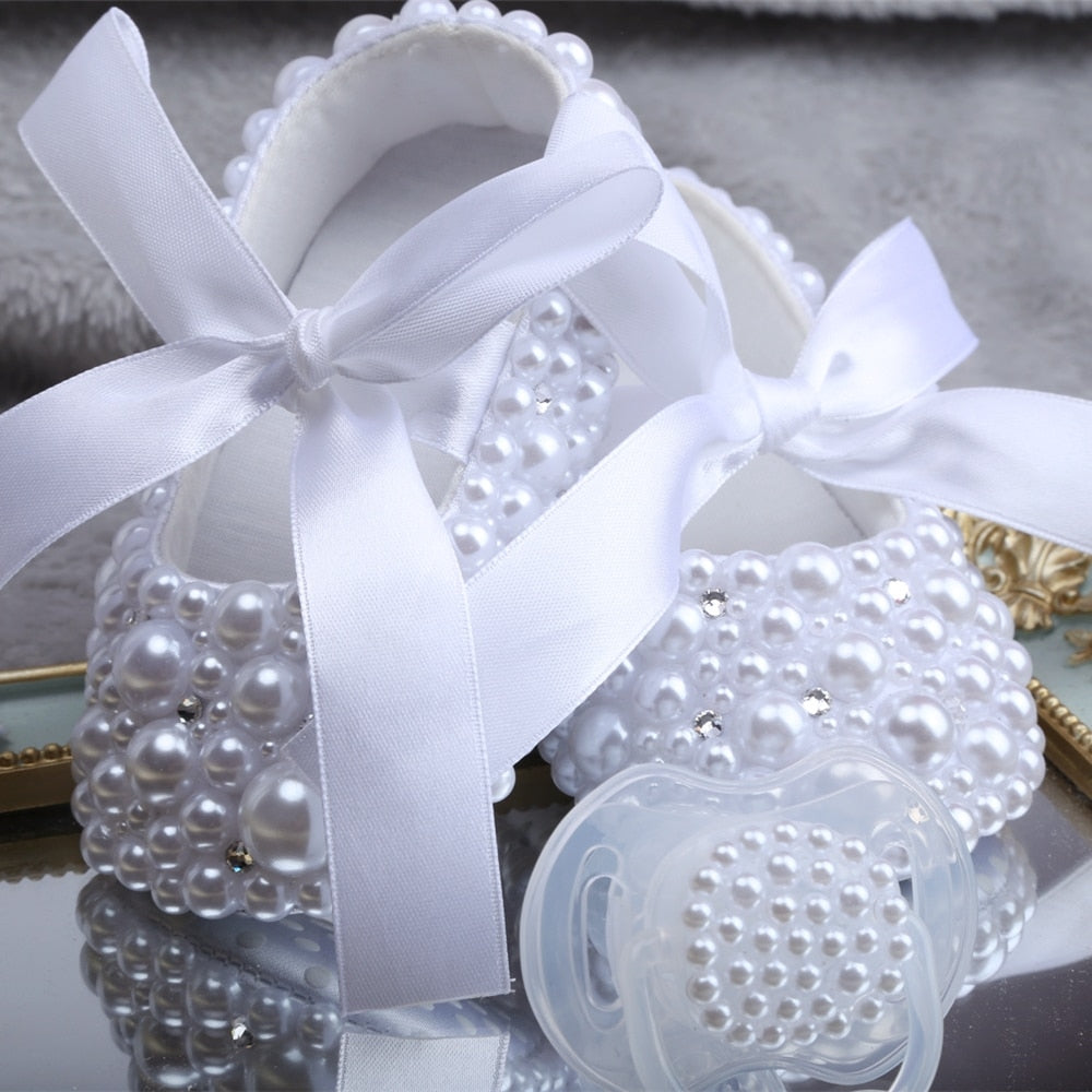 Elegant baby girl's white first walker shoes with delicate pearl and ribbon accents and pacifier.