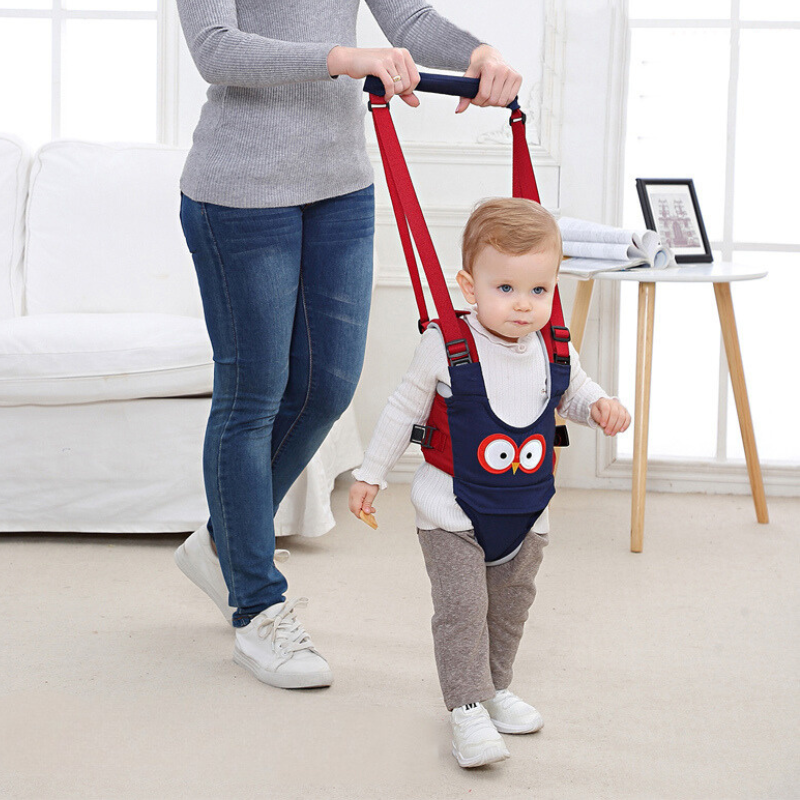 Baby in a blue walking harness with yellow straps, taking first steps with an adult's help.