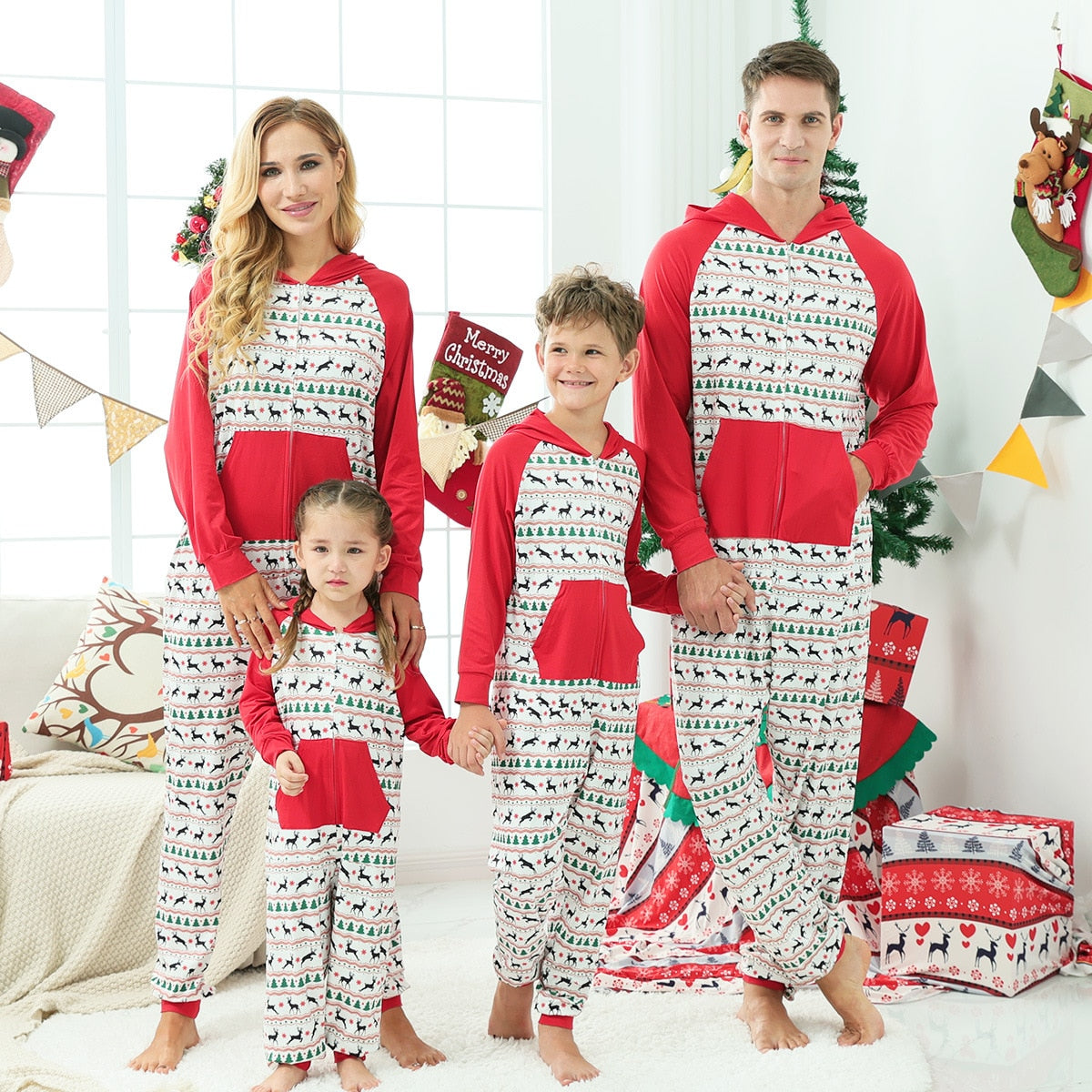 Family in matching red Christmas tree print one-piece rompers, smiling by a fireplace.
