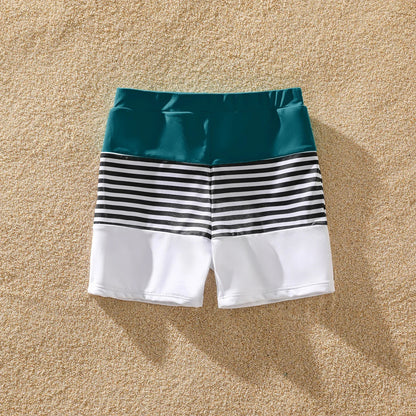 Family posing on a beach, all in green one-shoulder swimsuits & striped trunks