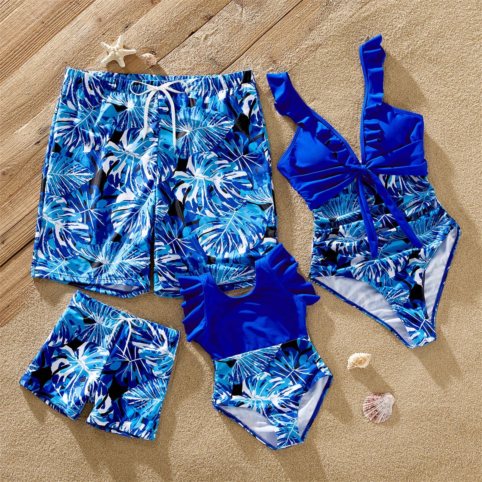 Family matching tropical leaf swimsuits: father in trunks, mother and girls in ruffle one-pieces, beach.