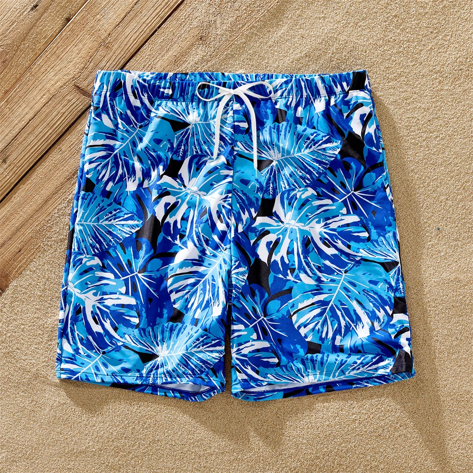 Dad in tropical leaf swim trunks, mom & daughter in matching ruffle one-piece swimsuits on sand.