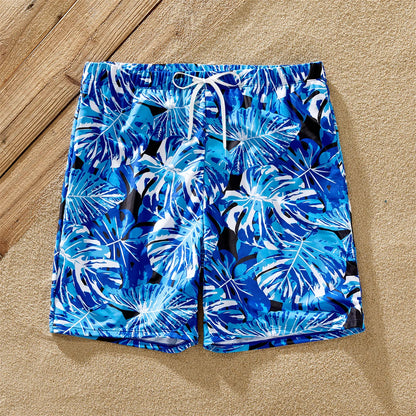 Dad in tropical leaf swim trunks, mom & daughter in matching ruffle one-piece swimsuits on sand.