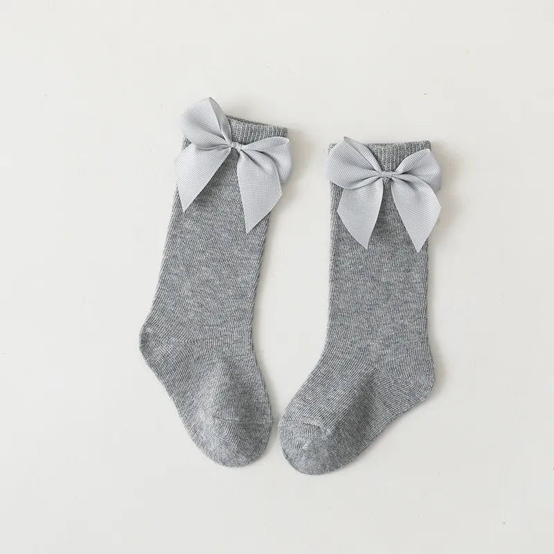 Folded pair of white cotton knee-high bow socks on a wooden surface; toddler essentials