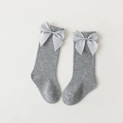 Folded pair of white cotton knee-high bow socks on a wooden surface; toddler essentials