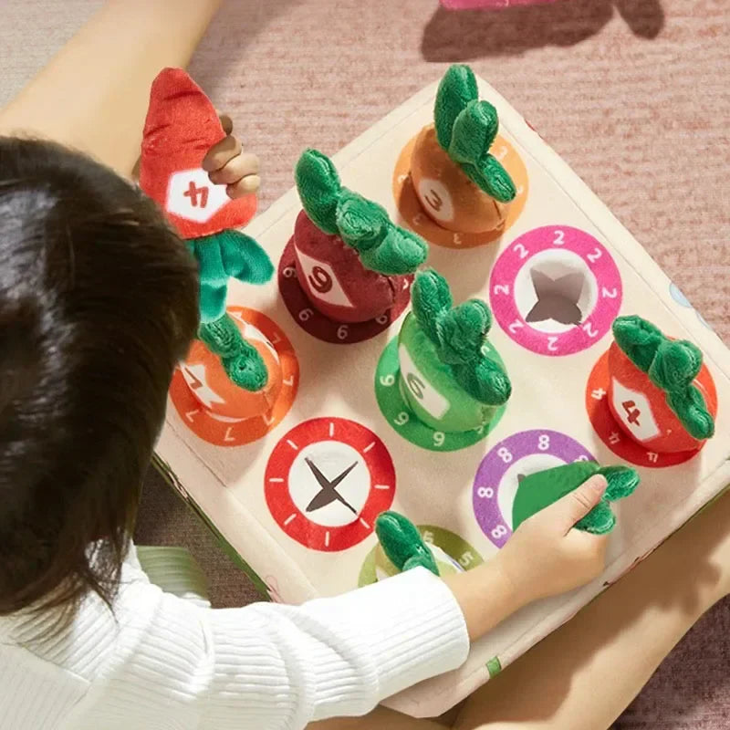 Child's hand reaching to pull a plush orange carrot from a green fabric hole on a sensory toy board.