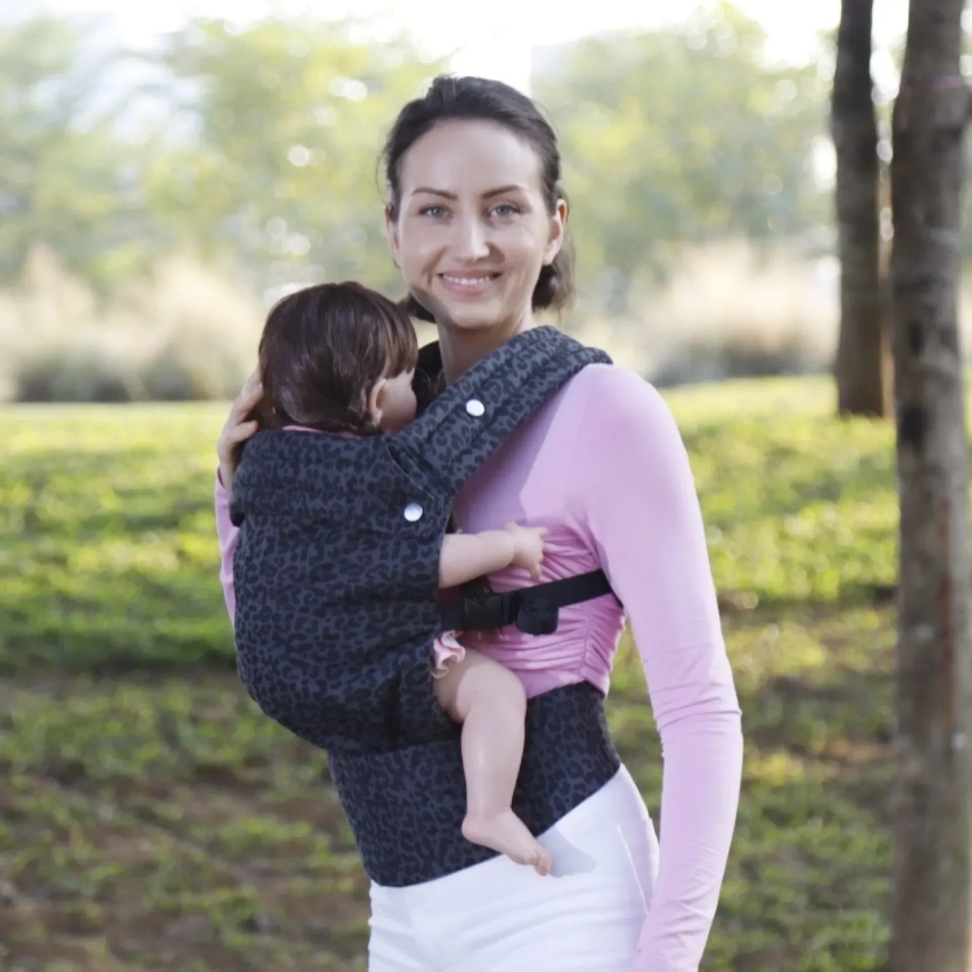 A parent smiling while wearing their baby in a gray, adjustable Mei Tai carrier